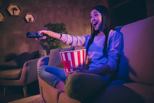 Photo of homey funny chinese lady good mood holding tv remote controller change channels eat popcorn movie night sit cosy couch casual outfit dark living room indoors