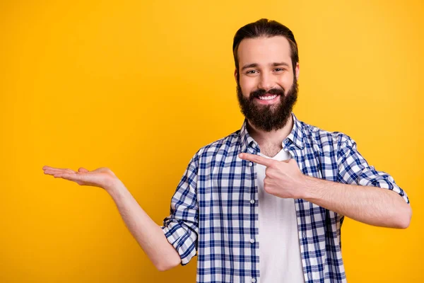 Ritratto di bello attraente allegro allegro contenuto fiducioso ragazzo barbuto in camicia a quadretti mostrando tenendo oggetto invisibile sul palmo isolato su brillante brillantezza vivace colore giallo sfondo — Foto Stock