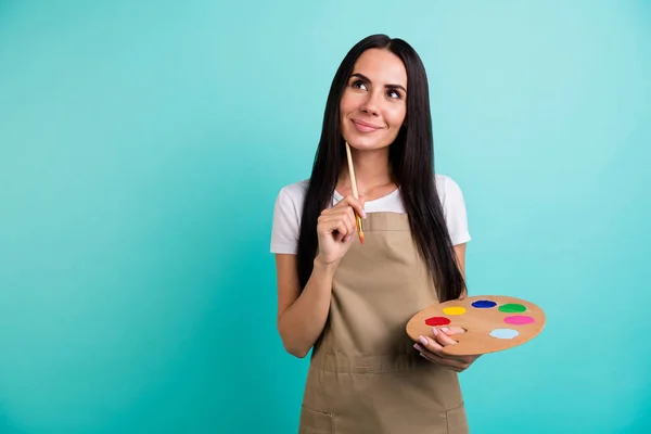 Foto de alegre positiva linda mujer beautfiul mirando hacia arriba en un ajuste de pensamientos fantaseando qué pintar sosteniendo pincel colorido paleta aislado color verde azulado fondo vivo —  Fotos de Stock