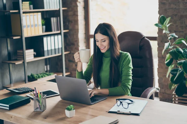 Profilkép csinos fiatal üzletasszony keres képernyő notebook asztal modern felhasználó chat partnerek inni forró tea bögre ül főnök szék viselni zöld garbó modern belső iroda — Stock Fotó