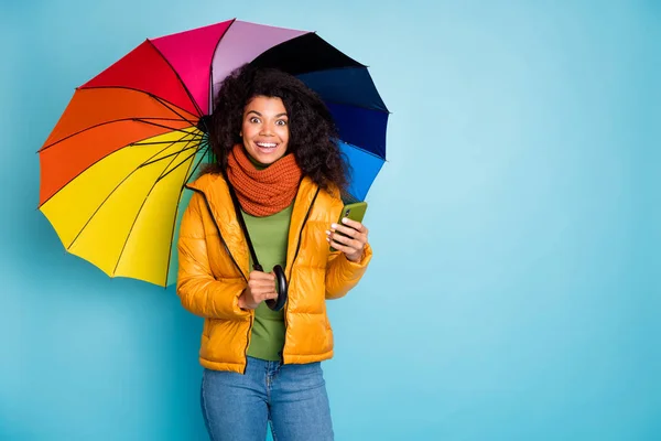Renkli şemsiyeli, koyu tenli bir kadının fotoğrafı. Modern sosyal ağ blogcusu şık bir palto giyiyor. Yeşil kazak, mavi arkaplan. — Stok fotoğraf