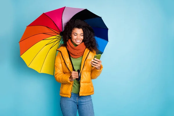 Güzel, koyu tenli bir kadının elinde renkli bir şemsiye, e-posta okuyor modern blogcu palto, yeşil kazak, eşarp, izole edilmiş mavi arka plan. — Stok fotoğraf