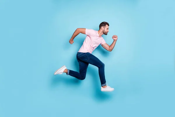 Volledige profiel foto van aantrekkelijke man springen hoge sportieve wedstrijden deelnemer dragen casual roze t-shirt jeans sneakers geïsoleerde blauwe kleur achtergrond — Stockfoto