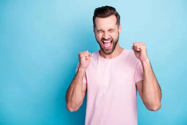 Foto de chico atractivo emociones extáticas ganar premio de dinero de la lotería gritando fuerte regocijo levantar los puños usar casual rosa camiseta aislado color azul fondo — Foto de Stock