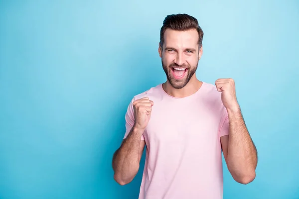 Photo of attractive guy ecstatic emotions win lottery money prize yelling loud rejoicing raise fists wear casual pink t-shirt isolated blue color background