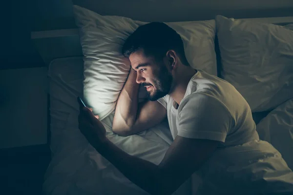 Topo acima do ângulo alto vista retrato do seu ele agradável atraente alegre alegre cara deitado na cama usando celular digital conversando à noite tarde à noite casa escuro iluminado quarto apartamento casa — Fotografia de Stock