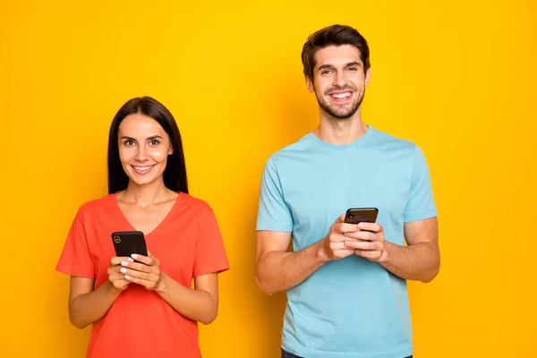 Foto de dos divertido chico señora personas pareja sostener teléfonos brazos lectura redes sociales post comentarios desgaste casual azul naranja camisetas aisladas color amarillo fondo —  Fotos de Stock
