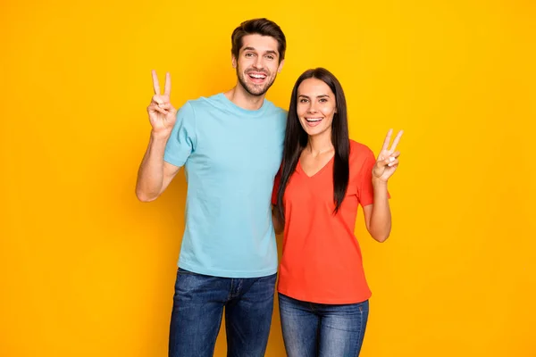 Φωτογραφία του funky δύο άτομα ζευγάρι τύπος και κυρία δείχνει σύμβολο v-σημάδι χαιρετισμό τους φίλους κατά τη συγκέντρωση κόμμα φορούν casual μπλε πορτοκαλί t-shirts τζιν απομονώνονται σε κίτρινο χρώμα φόντο — Φωτογραφία Αρχείου