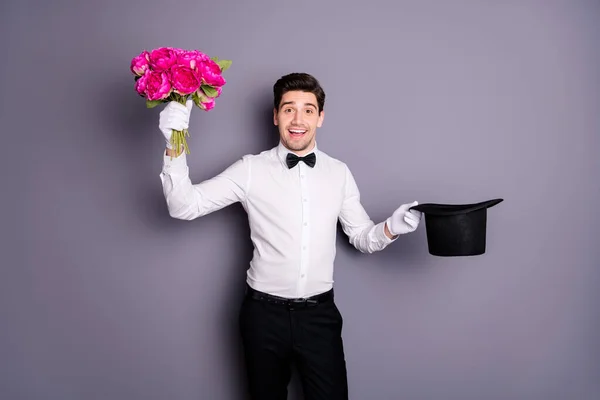 Des fleurs pour madame. Photo de beau spectacle homme tenir noir cylindre magicien obtenir pivoines fraîches tas de l'intérieur porter chemise blanche noeud papillon pantalon isolé couleur grise fond — Photo