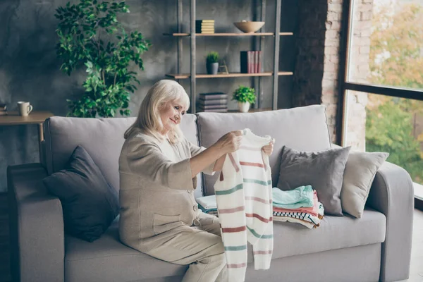 Profilo foto di stupefacente bianco dai capelli anziani nonna tenendo in ordine le mani pullover metterlo in pila primavera pulizia generale casa seduta divano soggiorno al chiuso — Foto Stock