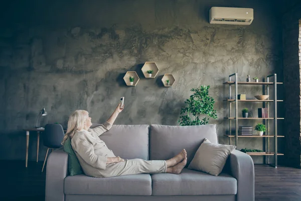 Profilo foto di affascinante bianco dai capelli anziani nonna cambiando modalità condizionatore accendere aria calda divano divano indossare abiti beige pastello soggiorno all'interno — Foto Stock