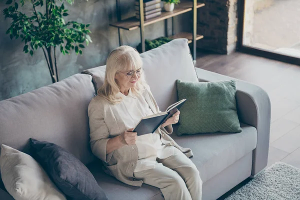 Фото блондинка очаровательный пожилой бабушки сидя удобный диван читая любимую книгу исторический роман хорошее свободное время носить бежевую пастельную одежду квартира гостиная в помещении — стоковое фото