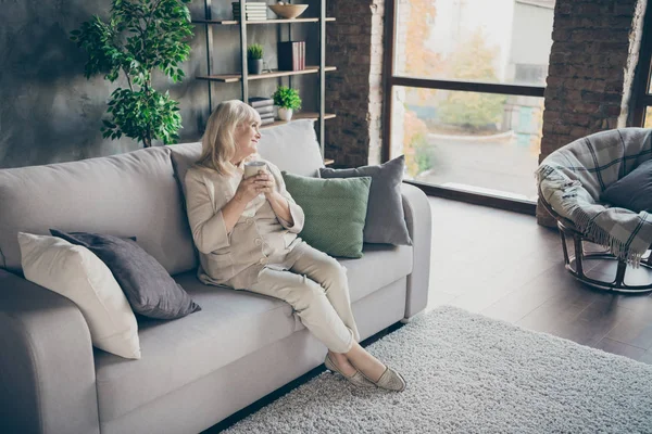 Bello attraente allegro sognante tipo dai capelli grigi bionda di mezza età nonna seduta sul divano a riposo bere tisana a loft industriale stile moderno casa appartamento appartamento — Foto Stock