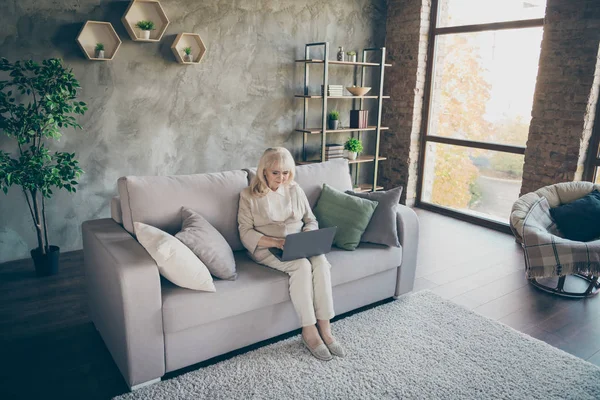 Nizza attraente concentrato dai capelli grigi concentrata bionda di mezza età nonna seduta sul divano a riposo utilizzando computer portatile chiamare i parenti a loft industriale stile moderno casa appartamento appartamento — Foto Stock