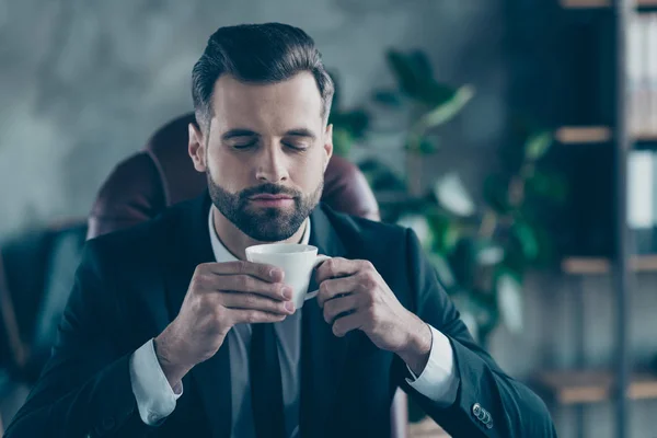 Photo of successful business guy hold coffee beverage mug thoughtful eyes closed enjoy pleasant smell wear black blazer shirt tie suit costume sitting chair office indoors — 스톡 사진