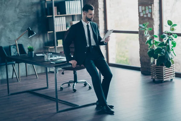 Foto de larga duración del exitoso hombre de negocios que espera reunión corporativa leer informes de lectores electrónicos mesa inclinada usar blazer negro camisa pantalones de corbata traje de pie oficina moderna en interiores — Foto de Stock