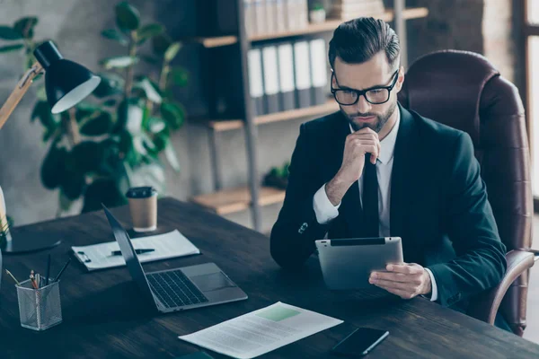 Foto av uppmärksam stilig affärskille fokuserad på att läsa e-reader rapport kontrollera ekonomi inkomstsiffror summor bära specs svart kavaj skjorta slips kostym sittstol kontor inomhus — Stockfoto