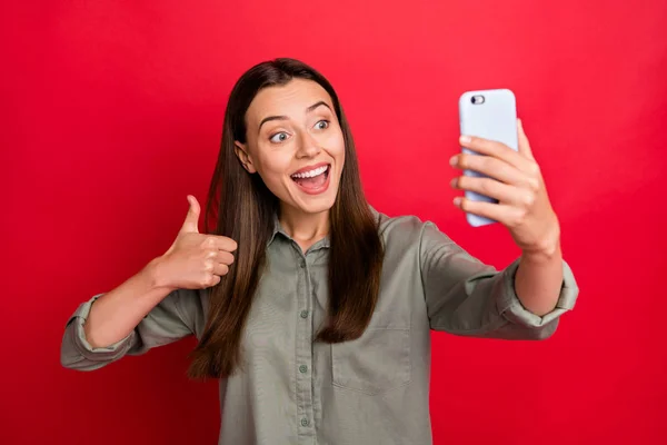 Фотографія молодої леді, що тримає телефон, робить селфі, піднімаючи великий палець, що виражає згоду розмовляти друзями неба, носять випадкову сіру зелену сорочку ізольовану червоний колір фону — стокове фото