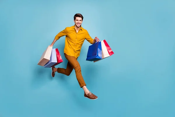 Foto de perfil de longitud completa de tipo divertido saltando alto corriendo rápido llevar muchas bolsas corriendo tienda siguiente tienda usar pantalones de camisa amarilla aislado fondo de color azul — Foto de Stock