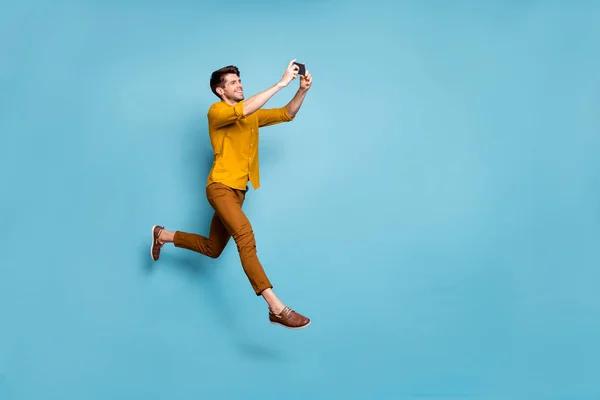 Full size profil fotografie legrační chlap skákání vysoké držení telefon dělat selfie on-line multimediální překlad nosit žluté košile kalhoty izolované modré barvy pozadí — Stock fotografie