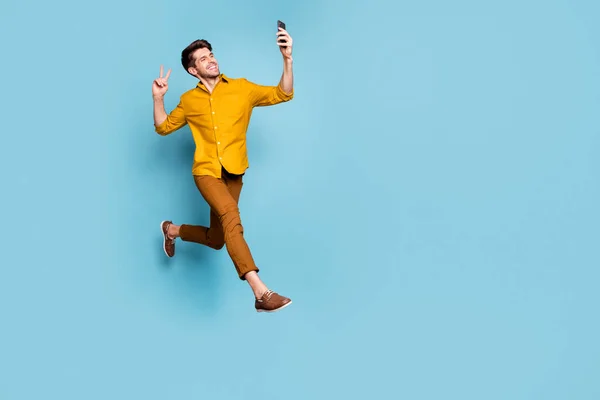 Full length photo of funny guy jumping high holding telephone making selfies showing v-sign symbol wear yellow shirt pants isolated blue color background