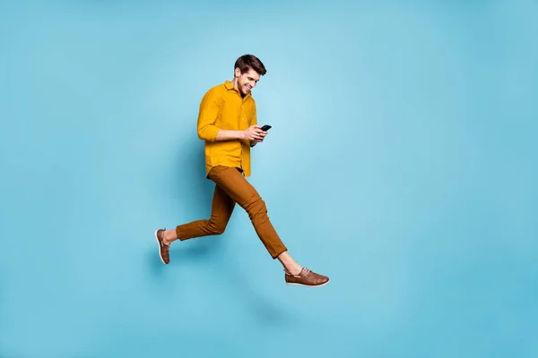 Volledige lengte profiel foto van grappige man springen hoge holding telefoon millennial schrijven e-mail lopen op straat dragen geel shirt broek geïsoleerde blauwe kleur achtergrond — Stockfoto