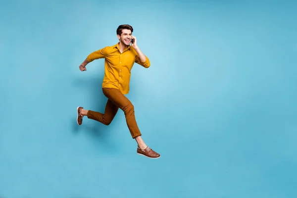 Jag är på väg. Full längd foto av galen kille hoppar högt rusa till fest innehav telefon talande vänner bära gul skjorta byxor isolerad blå färg bakgrund — Stockfoto