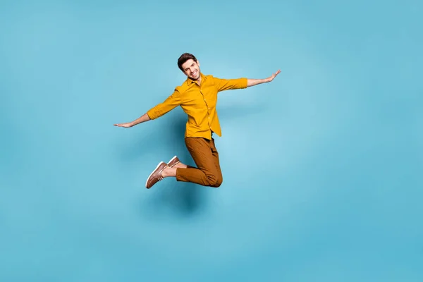 Foto de comprimento total de cara louco pulando alta segurando as mãos espalhadas por lados fingindo voo de pássaro usar calças camisa amarela isolado fundo de cor azul — Fotografia de Stock