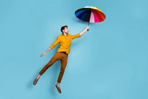 Foto in voller Länge von verrückten Kerl springen hoch halten farbige helle Regenschirm fliegen höher tragen gelbe Hemdhosen isoliert blaue Farbe Hintergrund — Stockfoto
