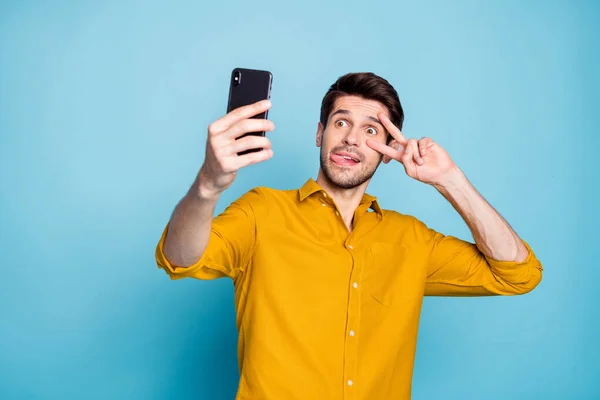 Foto di sciocco uomo ridicolo mostrando v-segno bastone toungue fuori tenendo telefono prendendo selfie comportarsi infantile isolato su sfondo di colore blu pastello — Foto Stock