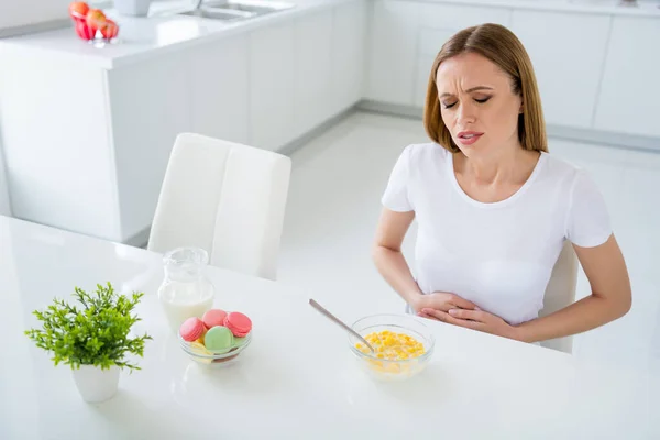 Foto de dona de casa muito chateada segurando barriga sofrendo dor terrível não pode comer leite café da manhã flocos de milho têm estômago chateado sentado mesa cozinha luz branca dentro de casa — Fotografia de Stock