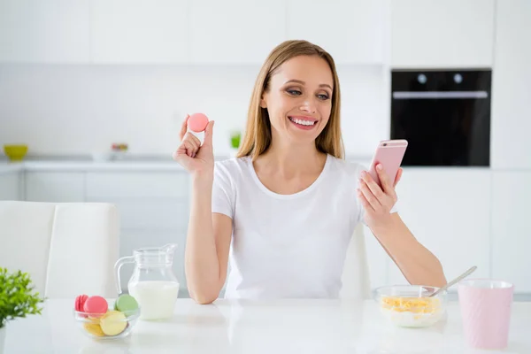 Foto de dona de casa bonita segurando telefone conversando com amigos comendo macaroons coloridos granola café da manhã leite na mesa cozinha luz branca dentro de casa — Fotografia de Stock