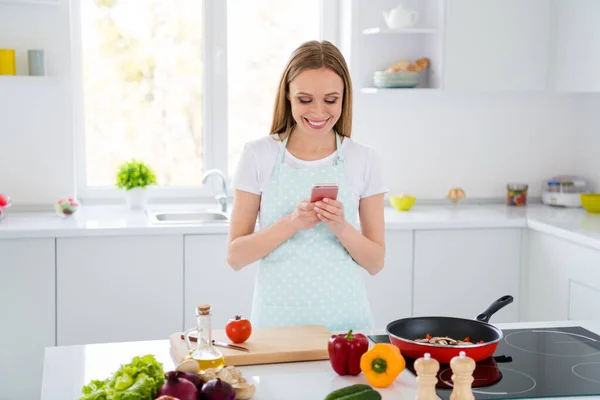 Foto de ama de casa fin de semana cocina de la mañana celebración de teléfono de pie cerca de la comida sartén lectura influencer en línea cocinar blog cocina blanca en el interior — Foto de Stock