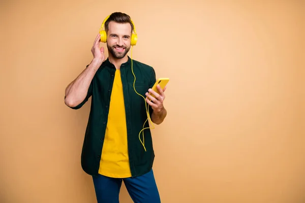 Фото привабливого привабливого хлопця-гіпстера, який тримає телефонні руки, слухаючи популярні пісні прохолодні сучасні жовті навушники, носять повсякденне вбрання ізольований бежевий кольоровий фон — стокове фото