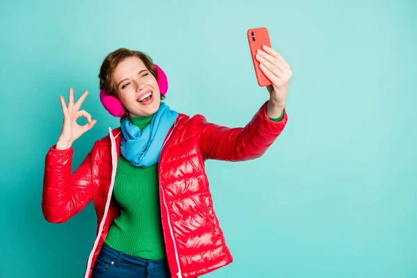 In Ordnung. Foto von lustigen hübschen Dame halten Telefon machen Selfies zwinkernden Auge zeigt okay Symbol tragen roten Mantel Schal rosa Ohrenschützer Pullover Hose isoliert Krickente Farbe Hintergrund — Stockfoto