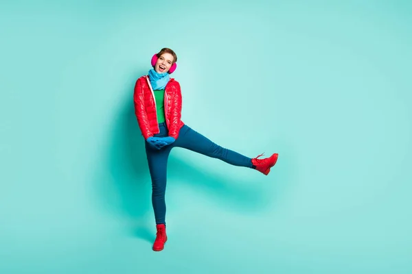 Komik bayan kış günü yürüyüş sokağının tam boy fotoğrafı en fazla boş zamanı bacağını kaldırarak geçirir günlük kırmızı palto eldiven giyer, pembe kulaklık pantolon, izole edilmiş deniz mavisi arka plan. — Stok fotoğraf
