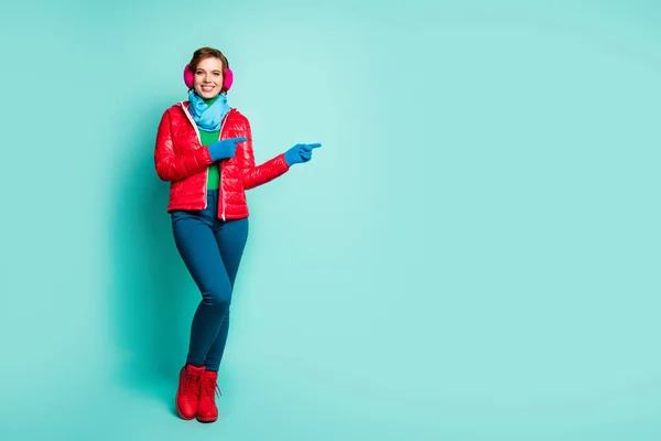 Full body photo of amazing lady directing fingers empty space offer sale prices season shopping wear red overcoat blue scarf pink ear muffs pants boots isolated teal color background