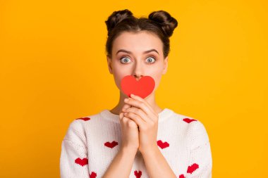 Closeup photo of pretty funny lady hold little paper heart hiding mouth postcard shy person wear white red hearts pattern pullover isolated yellow color background clipart