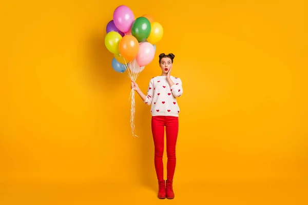 Foto de tamaño completo de la señora divertida celebrar globos de aire de colores boca abierta inesperada sorpresa usar corazones patrón suéter pantalones rojos zapatos aislados color amarillo fondo — Foto de Stock