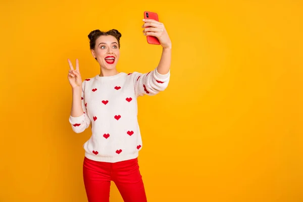 Photo of funny pretty lady hold telephone hands taking selfies showing v-sign symbol for online blog wear hearts pattern white sweater red pants isolated yellow color background