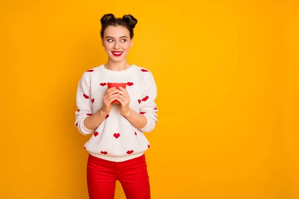 Photo of beautiful nice lady holding hot coffee beverage cup toothy smiling look side empty space wear hearts pattern white sweater red pants isolated yellow color background — 스톡 사진