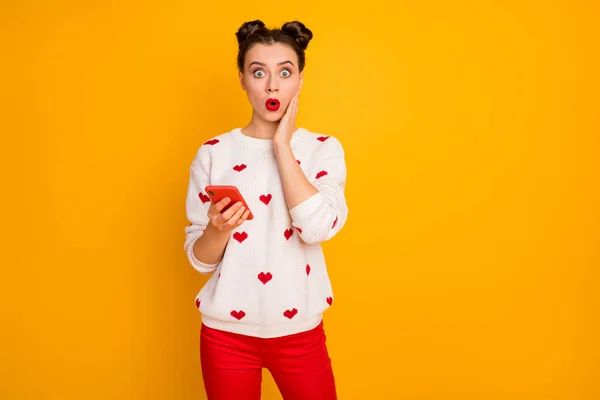 Фото досить шокованої леді тримає телефон читати неймовірно погані погані новини руки на щоці носити серця візерунок білий пуловер червоні штани ізольовані жовтий колір фону — стокове фото