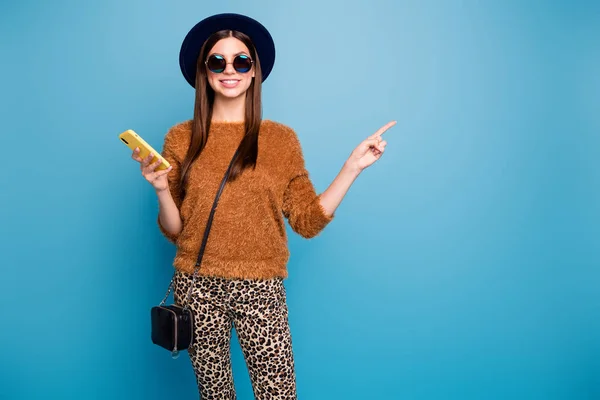 Positivo alegre estudiante chica uso smartphone repost anuncios promoción punto dedo copyspace desgaste retro sombrero bolso marrón suéter casual pantalones aislados sobre azul color fondo — Foto de Stock