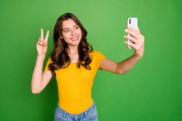 Фото гарненької кучерявої леді, що тримає телефони, що показують символ v-знака, кажуть, що привіт носить звичайні жовті футболки джинси ізольовані яскраво-зеленого кольору фону — стокове фото