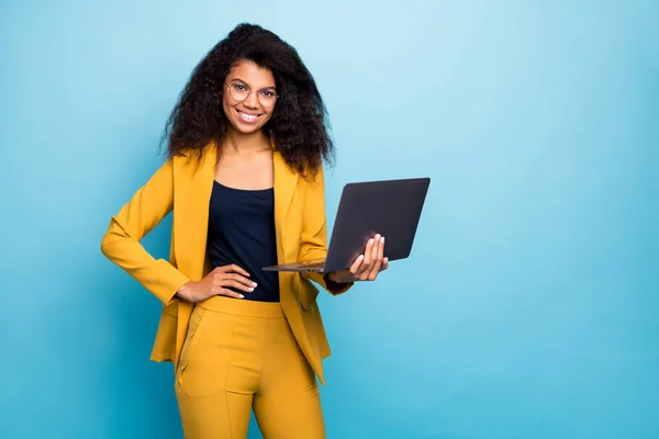 Foto de bonita asistente de piel oscura dama ondulada sosteniendo portátil información de búsqueda ayuda a internet jefe amigable trabajador usar especificaciones traje amarillo pantalones de chaqueta aislado color azul fondo —  Fotos de Stock