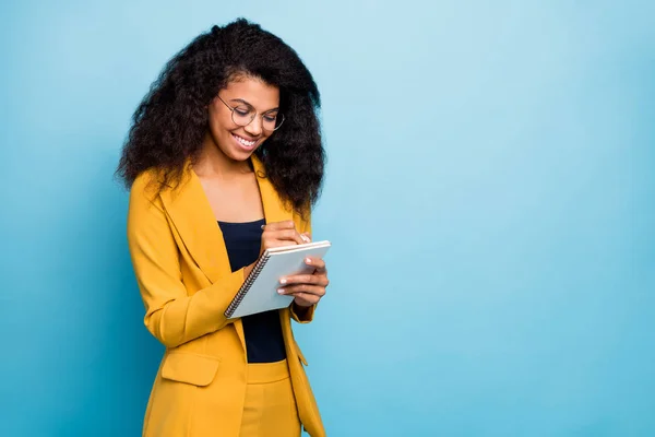 Foto van mooie donkere huid business dame creatieve schrijver opmerkend leuke gedachten op papier planner schrijven to-do lijst te dragen specs stijlvolle geel kantoor pak geïsoleerde blauwe kleur achtergrond — Stockfoto