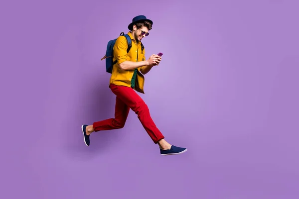 Perfil em tamanho completo foto lateral do cara hipster positivo relaxar viagem salto uso smartphone pesquisa descontos correr rápido desgaste camisa amarela calças vermelhas mochila isolado cor violeta fundo — Fotografia de Stock