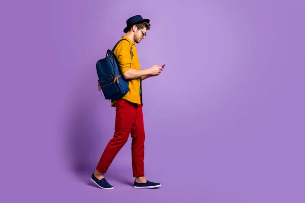 Full size profiel kant van geconcentreerde jongen lopen ga naar de universiteit gebruik smartphone teksttype social media sms-berichten dragen geel shirt blauw retro hoofddeksel rugzak geïsoleerde violet kleur achtergrond — Stockfoto