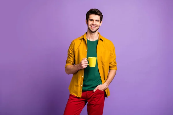 Foto de homem positivo alegre com cerda segurando mão no bolso de calças sorrindo toothily segurando xícara de café isolado cor pastel violeta fundo — Fotografia de Stock