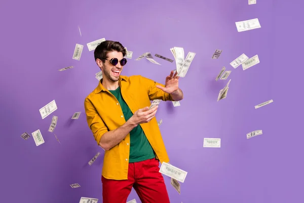 Foto de alegre emocionado hombre blanco de moda tirando dinero en efectivo mostrando su riqueza sonriendo toothily en pantalones rojos aislados color violeta fondo — Foto de Stock